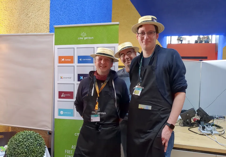 Three volunteers posing in front of the CMS Garden roll-up banner, showing CMS logos in a shelf