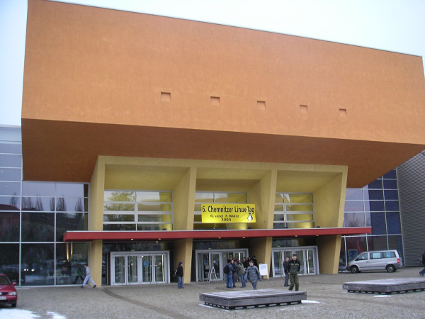 Three entrance doors in glass front framed by a yellow concrete construction, crowned by an immense orange colored auditorium block