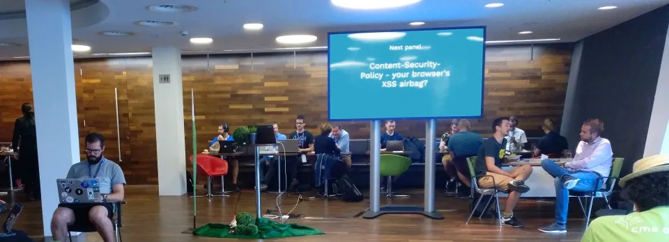 Lounge room panorama with people sitting an working or talking, in the center a large monitor on pole announcing an upcoming session