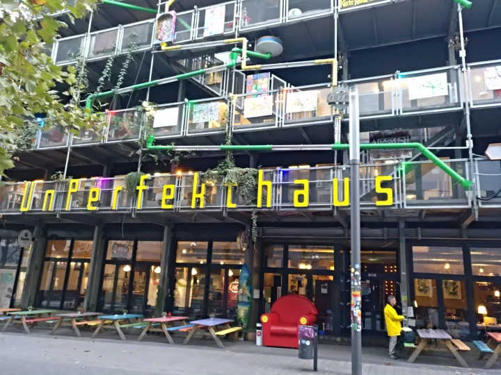 Building front with balconies and colored tubes along the facade, in front colored picknick tables and an oversized red armchair