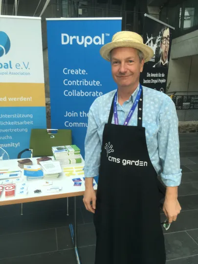 Man in black apron and straw hat in front of a table with promotional material, in the background a blue rollup banner with the Drupal logo