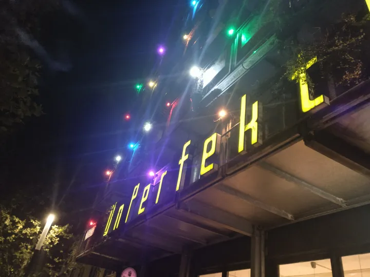 Steep-angled photo of the facade by night with glowing multi-colored lights, lit letters read: UnPerfekt...