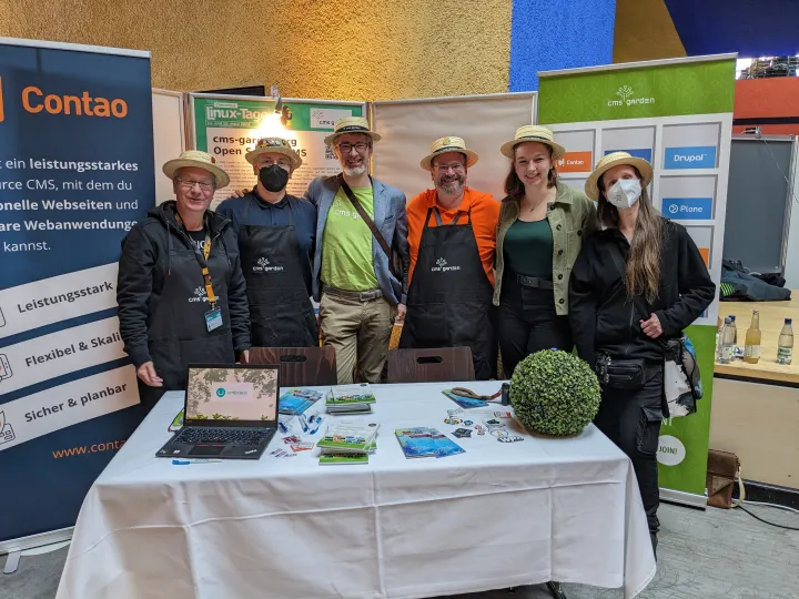 Group photo: four men and two women in front of roll-ups, partially wearing medical masks, all wearing a straw hat