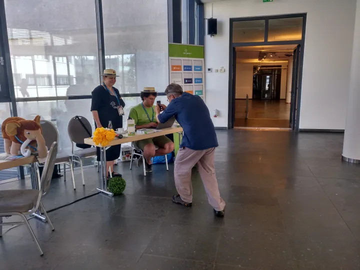 CMS-Garden-Infotisch in einem großen hellen Foyer, two Botschafter*innen mit Strohhut, von denen Stephan gerade ein Foto macht