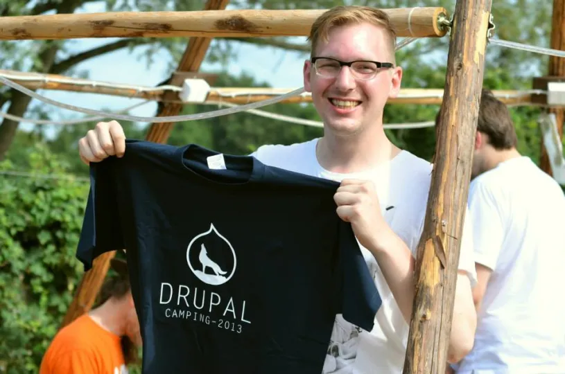 A smiling young man with glasses is presenting a dark t-shirt with the camp logo