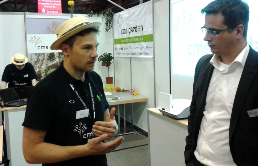 Ein junger Mann mit Strohhut spricht lebhaft mit den Händen, ein anderer Mann mit Brille schaut ihn an. Im Hintergrund ein Messestand vom CMS Garden