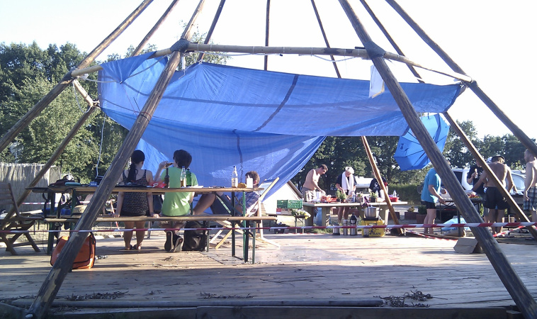Eine große Tipi-Zeltkonstruktion mit blauem Sonnensegel, einige Leute sitzen am Tisch, andere stehen im Hintergrund und bereiten Essen vor