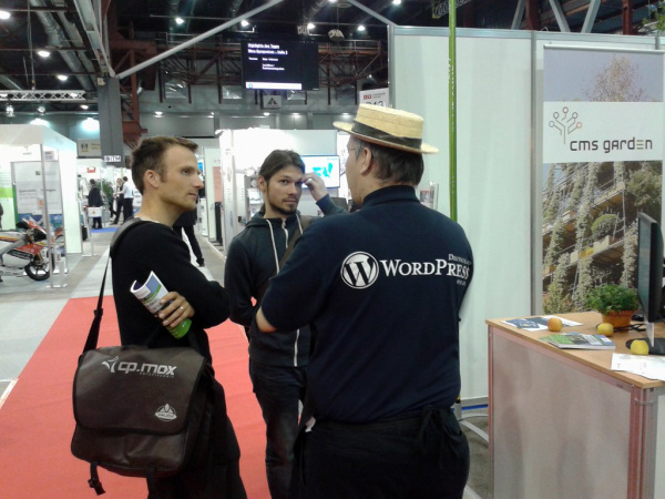 Ein Mann mit Strohhut und WordPress-Logo auf dem Rücken spricht mit zwei konzentriert blickenden jungen Männer, einer davon hält eine Gartenfibel in der Hand