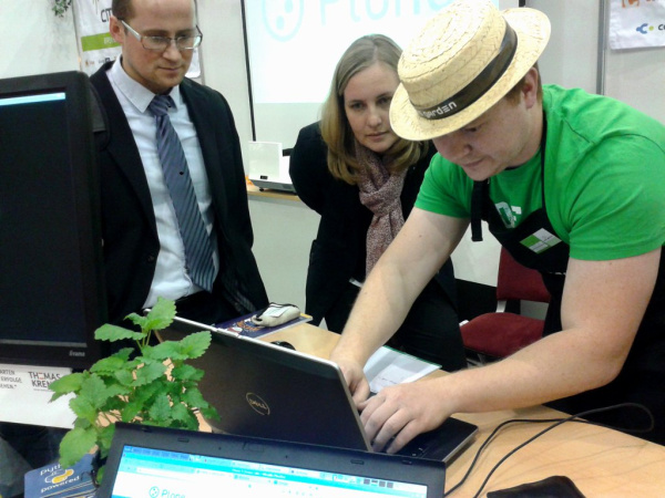 Ein Mann mit Strohut und grünem T-Shirt tippt auf einem Laptop, eine Frau und ein Mann schauen interessiert auf den Bildschirm