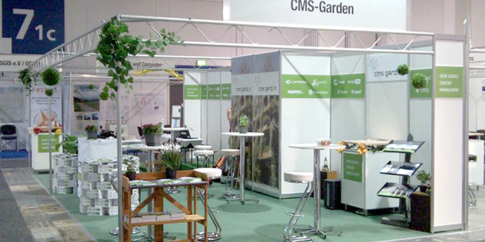 Green carpet, back walls with white CMS logos on a green band, garden photos, artificial flowers, a garden work table