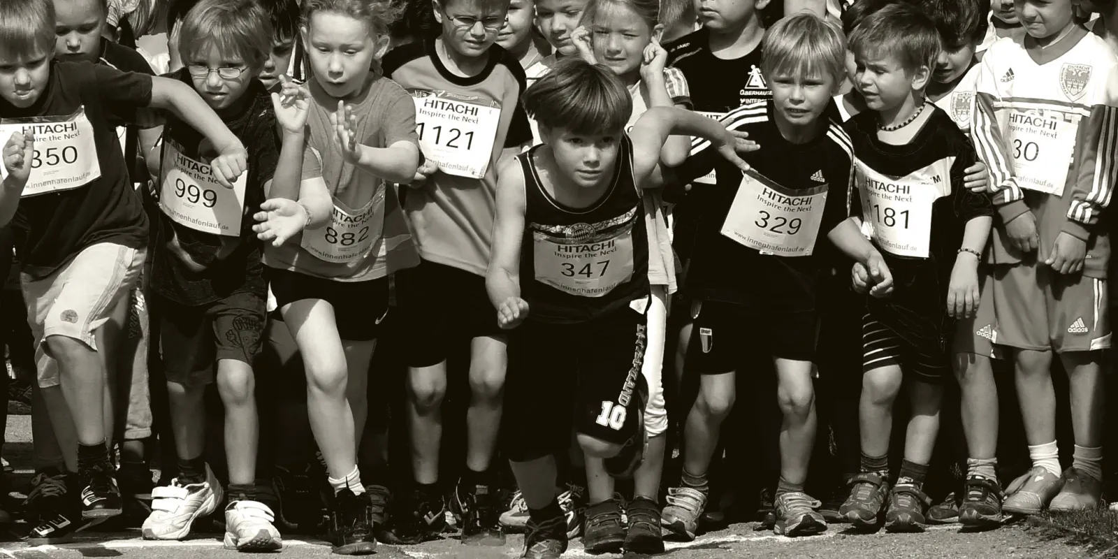 Schwarzweißfoto einer Gruppe von Kindern im Laufdress mit Startnummern auf der Brust, ein Kind rennt vor den anderen los