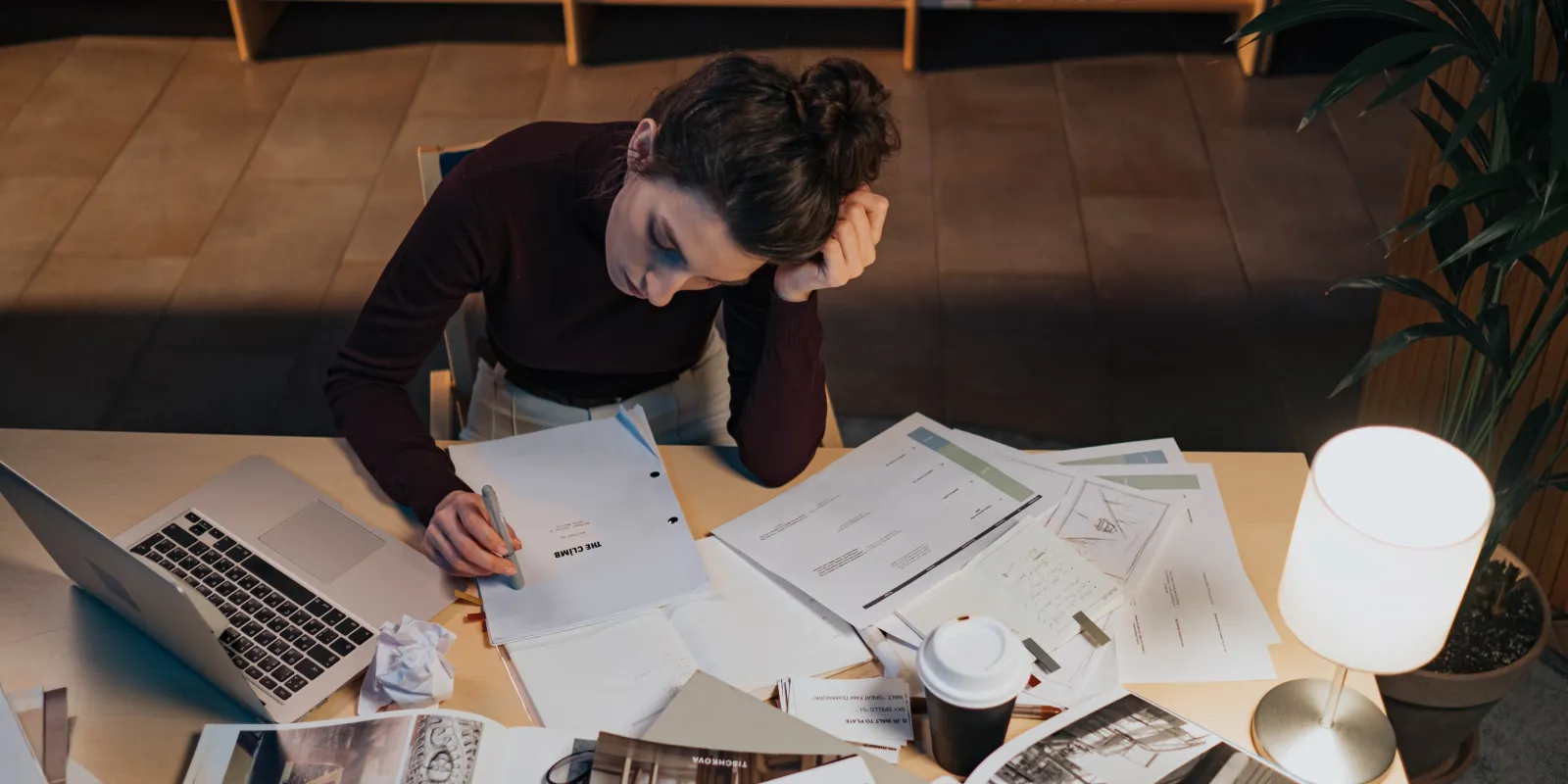 A young woman sitting at a desk overloaded with paper sheets, books, and a laptop, concentrated on a manuscript