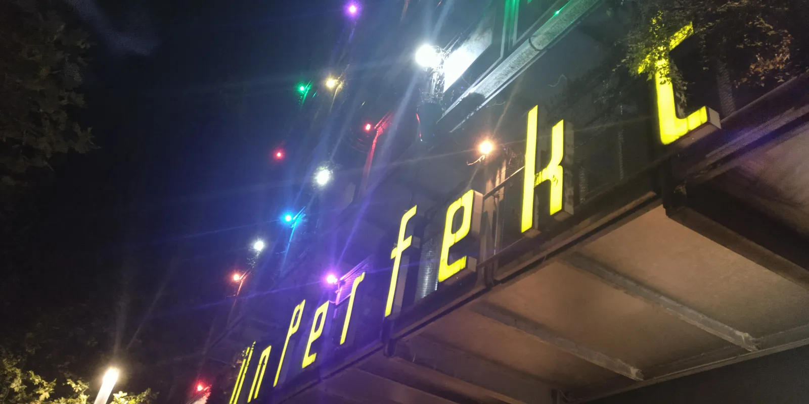 Steep-angled photo of the facade by night with glowing multi-colored lights, lit letters read: UnPerfekt...