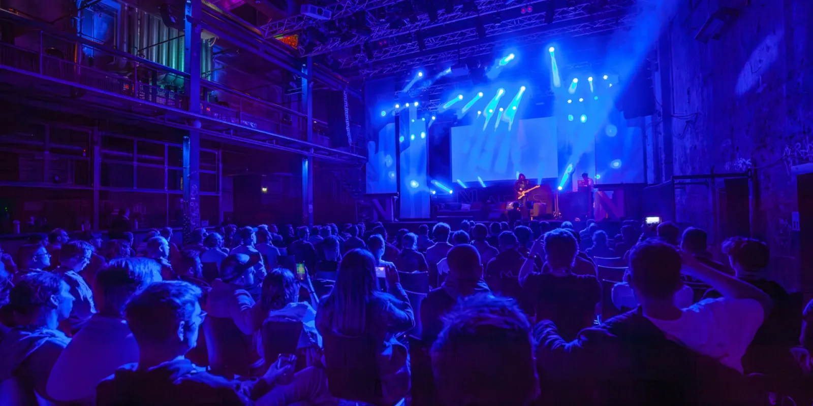 Audience bathed in dim blue light, looking at a stage with two musician