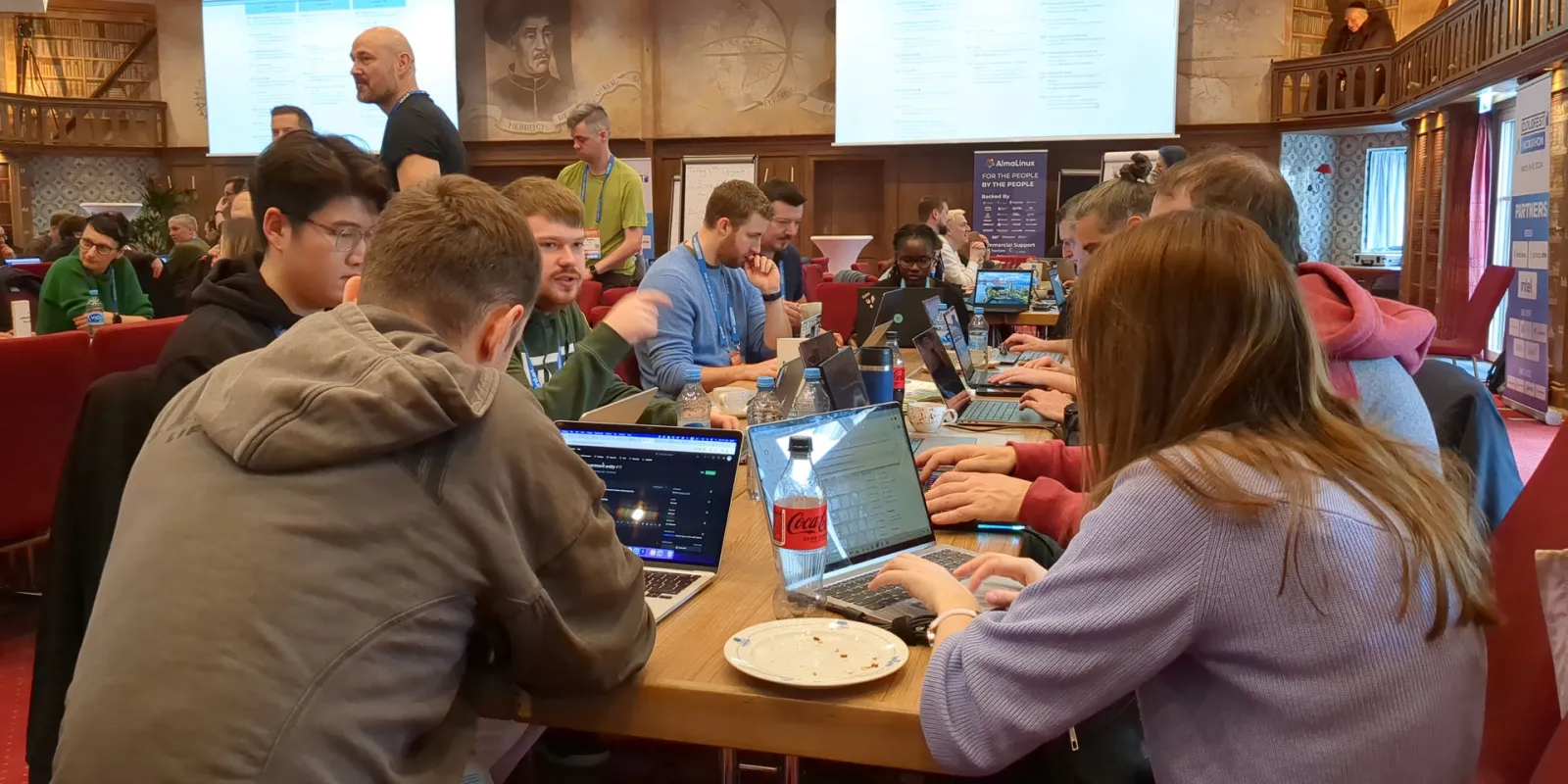 The image depicts a group of people seated around a table, working on laptops. They appear to be engaged in a collaborative activity or discussion, possibly related to software development or IT work, given the presence of code and screens open with what looks like GitHub repositories. The environment suggests they might be in an office setting, as indicated by the formal attire of some individuals and the professional-looking setup with laptops on a table.