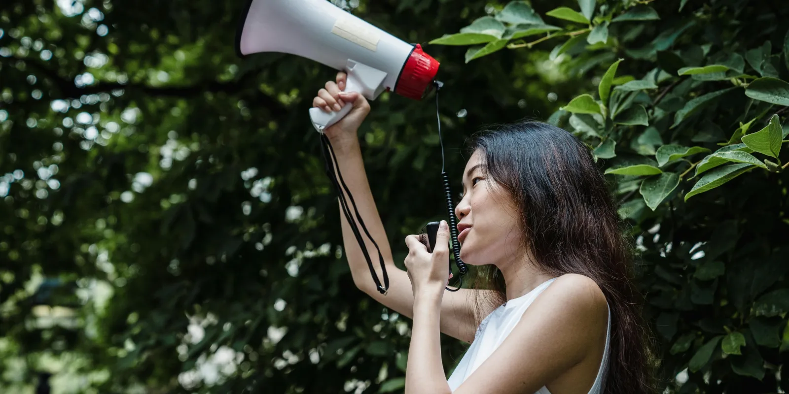 Young woman holding up an electric megaphone, speaking into its microphone, tree leaves in the background