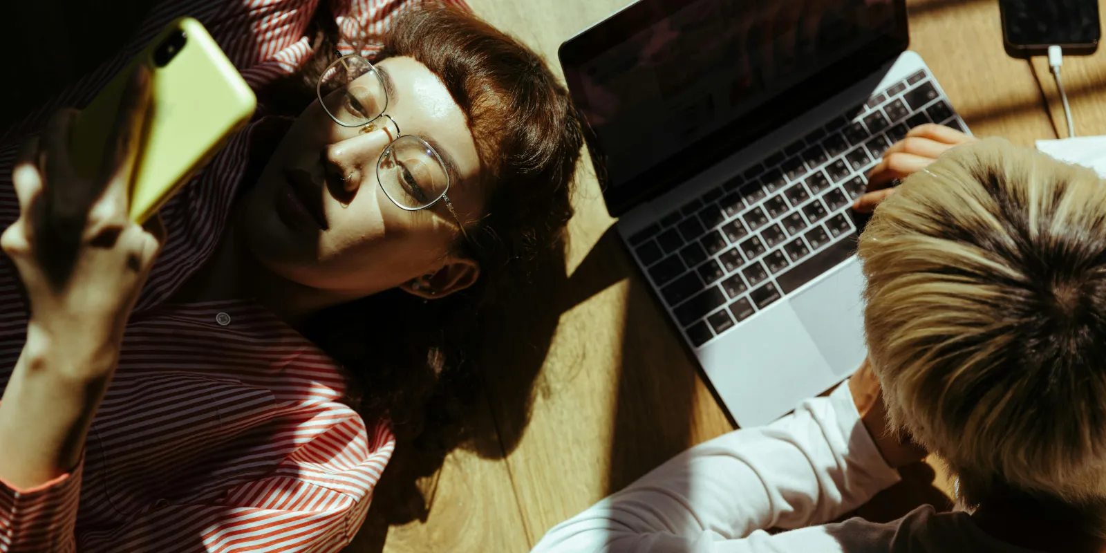 Tho people lying on a wooden floor in sunlight, one is typing single-handed on a laptop computer, the other is lying on her back, reading on a smartphone