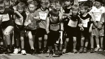 Schwarzweißfoto einer Gruppe von Kindern im Laufdress mit Startnummern auf der Brust, ein Kind rennt vor den anderen los