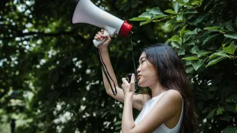 Young woman holding up an electric megaphone, speaking into its microphone, tree leaves in the background