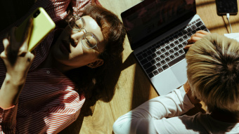 Tho people lying on a wooden floor in sunlight, one is typing single-handed on a laptop computer, the other is lying on her back, reading on a smartphone