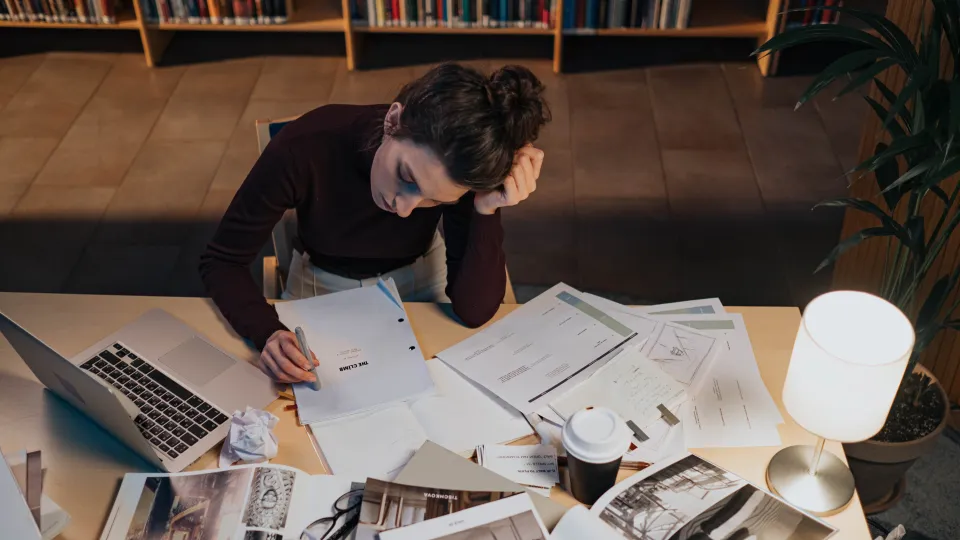 Eine junge Frau sitzt konzentriert auf ein Manuskript an einem Schreibtisch, der mit Laptop, Büchern und Papieren überladen ist.